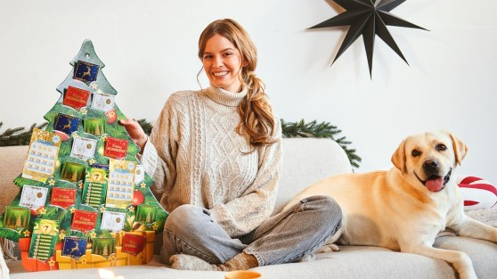 Frau sitzt neben Hund auf Couch und hält Rubbellos-Adventskalender in Tannebaum-Design | WestLotto